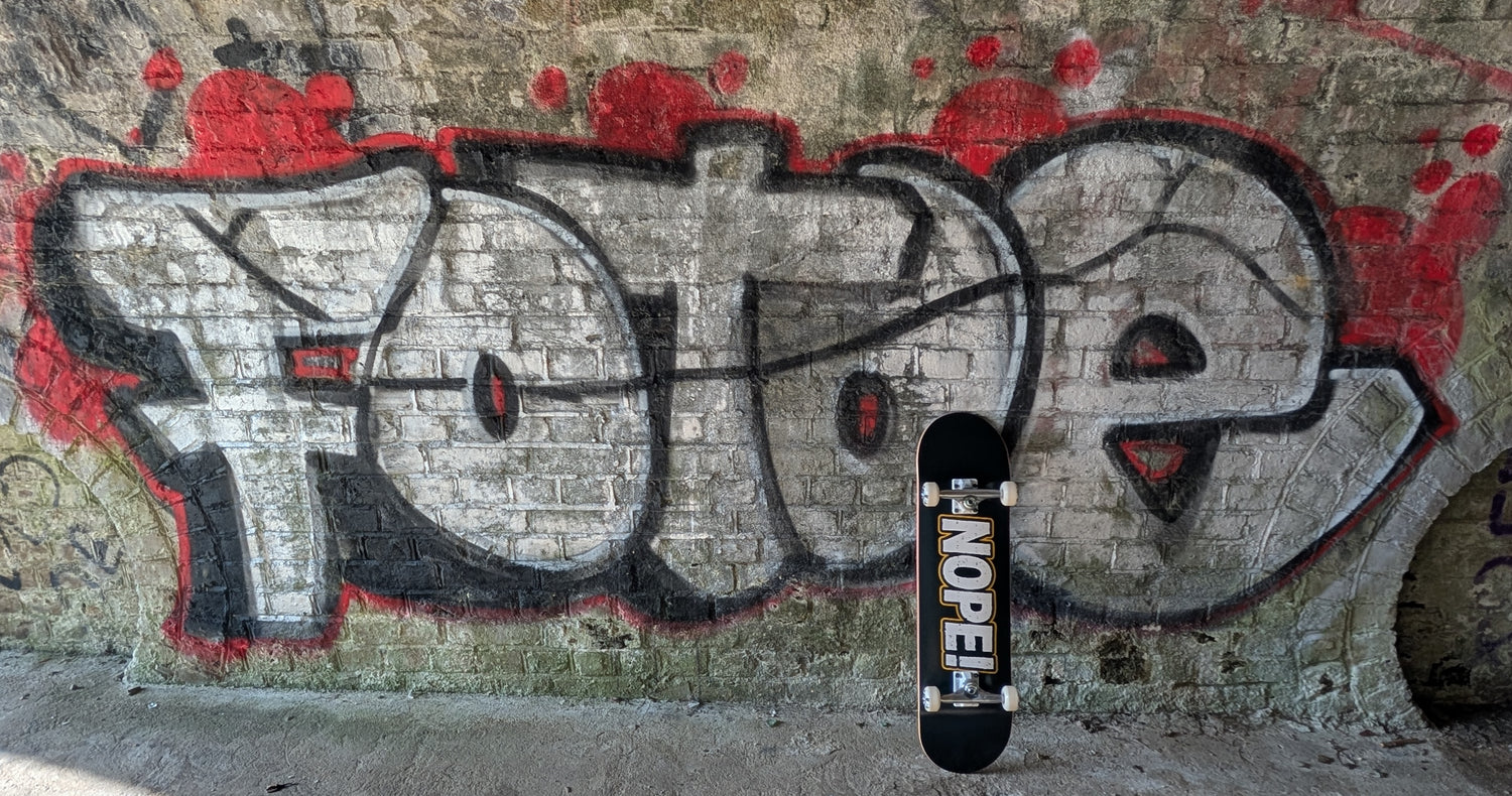 A skateboard with a black deck and white wheels leans against a brick wall covered in graffiti, with the word 'FOXE' painted behind it.