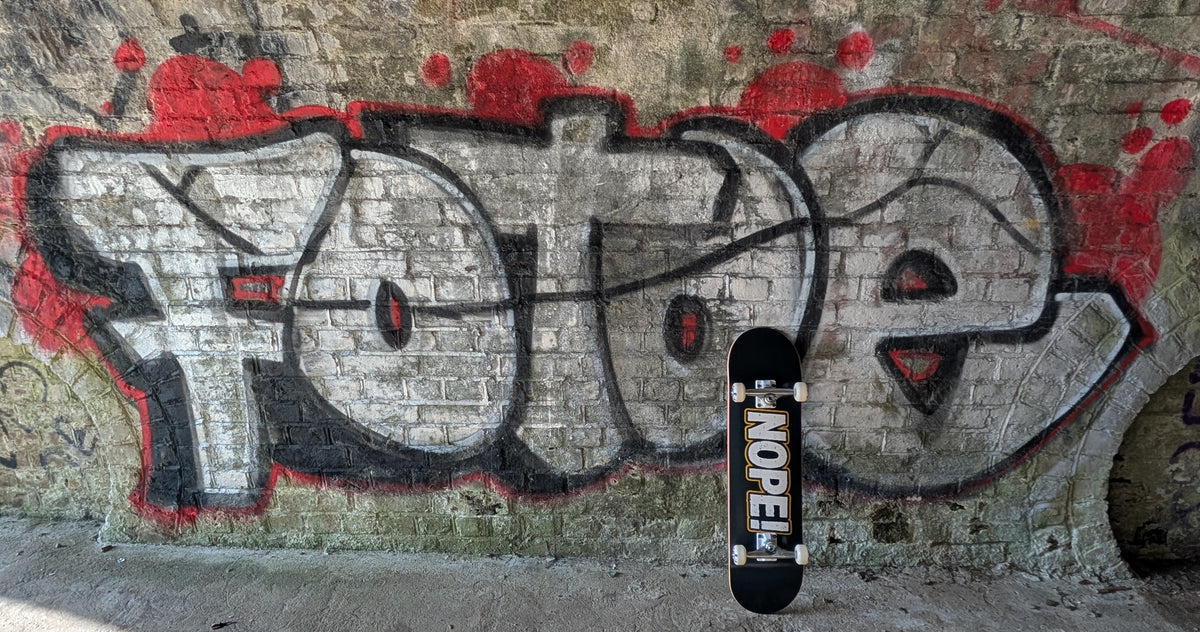 A skateboard with a black deck and white wheels leans against a brick wall covered in graffiti, with the word 'FOXE' painted behind it.